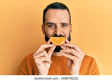 Young Man With Beard Holding Orange Slice On Mouth As Funny Smile Looking Positive And Happy Standing And Smiling With A Confident Smile Showing Teeth 
