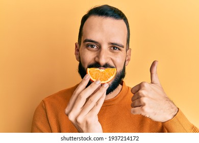Young Man With Beard Holding Orange Slice On Mouth As Funny Smile Smiling Happy And Positive, Thumb Up Doing Excellent And Approval Sign 