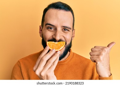 Young Man With Beard Holding Orange Slice On Mouth As Funny Smile Pointing Thumb Up To The Side Smiling Happy With Open Mouth 