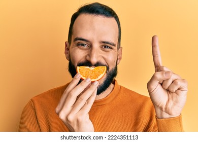 Young Man With Beard Holding Orange Slice On Mouth As Funny Smile Smiling With An Idea Or Question Pointing Finger With Happy Face, Number One 