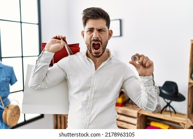 Young Man With Beard Holding Shopping Bags And Bitcoin Angry And Mad Screaming Frustrated And Furious, Shouting With Anger. Rage And Aggressive Concept. 