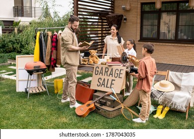 Young Man With Beard Looking Through Book While Choosing It At Yard Sale, Woman And Her Kids Waiting For Customers Answer