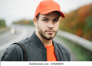 Young man with beard on his serious sexy handsome face in dark casual jacket and red baseball cap on head on background of road way outdoor