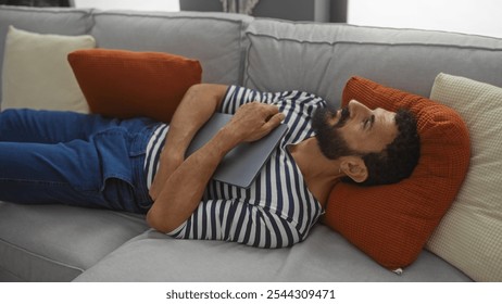 Young man with beard relaxing on a couch in a living room holding a tablet, with striped shirt and pillows around in a cozy indoor setting - Powered by Shutterstock