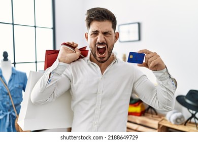 Young Man With Beard At Retail Shop Holding Shopping Bags And Credit Card Angry And Mad Screaming Frustrated And Furious, Shouting With Anger. Rage And Aggressive Concept. 