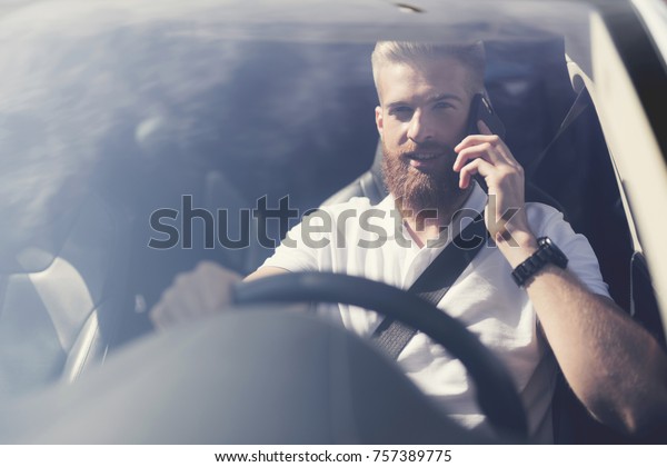 A young man with a\
beard sits at the wheel of an electric vehicle. He feels confident\
in the cabin of this car. He is wearing a seat belt. He is talking\
on the phone.