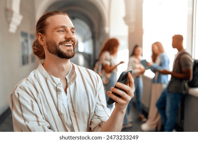 A young man with a beard is smiling while using a smartphone in a bright university hallway. In the background, a group of friends can be seen, indicating a fun and casual atmosphere among students