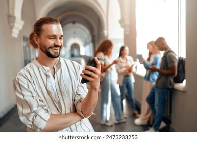 A young man with a beard is smiling while using a smartphone in a bright university hallway. In the background, a group of friends can be seen, indicating a fun and casual atmosphere among students
