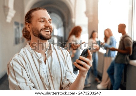 A young man with a beard is smiling while using a smartphone in a bright university hallway. In the background, a group of friends can be seen, indicating a fun and casual atmosphere among students