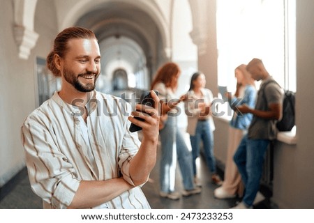 A young man with a beard is smiling while using a smartphone in a bright university hallway. In the background, a group of friends can be seen, indicating a fun and casual atmosphere among students