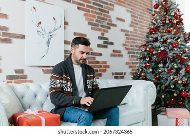 Young Man With A Beard Smiling And Working On A Laptop From Home, Sitting On The Couch During The New Year Holidays