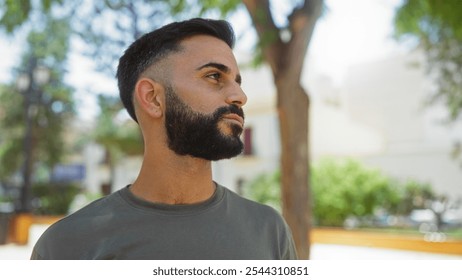 Young man with a beard standing outdoors in a vibrant urban park on a sunny day, showcasing a calm and thoughtful expression - Powered by Shutterstock