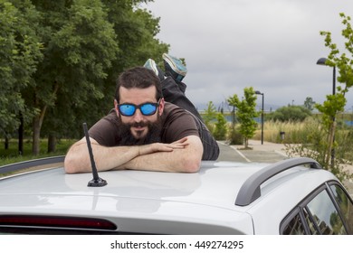 Young Man With Beard And Sunglasses Lying On The Roof Of A Car.