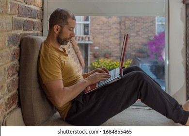 Young Man With Beard, Sweat Pants, T-shirt And His Laptop Over The Legs Working At Home By A Window. Home Office Concept