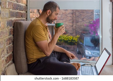 Young Man With Beard, Sweat Pants And T-shirt Working At Home By A Window, Having A Hot Drink While Looking At The Screen Of His Laptop