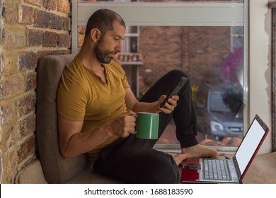 Young Man With Beard, Sweat Pants And T-shirt Working At Home Having A Hot Drink While Checking His Smartphone