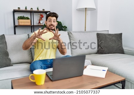 Young man with beard using laptop at home afraid and terrified with fear expression stop gesture with hands, shouting in shock. panic concept. 