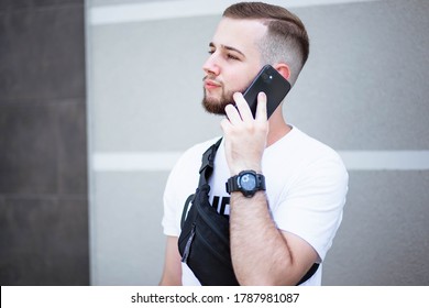 Young man with a beard walking and talking on a cell phone. Good talk. Happy young bearded man talking on the mobile phone and looking away while standing near the grey wall. Video chat. Phone.