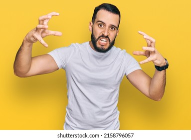 Young man with beard wearing casual white t shirt shouting frustrated with rage, hands trying to strangle, yelling mad 