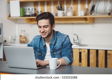 Young Man With Beard Working On Laptop. Happy Guy Is Sitting In The Kitchen And Chatting With His Friends