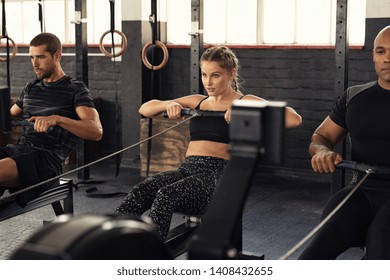 Young Man And Beautiful Woman Working Out With Rowing Machine At Crossfit Gym. Athletic Class Doing Exercise With Rowing Machine. Group Of Fitness Concentrated People In Sportswear Training.