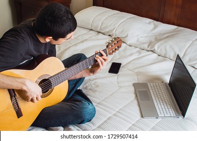 Young Man In A Bed Playing Guitar And Using A Computer To Learn At Home. Leisure At Home: Music And Technology, Learn Guitar With Online Classes.