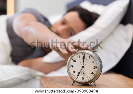 Young man being awakened by an alarm clock in his bedroom. Happy wake up of a man lying on the bed and stopping alarm clock. Man snoozing the alarm clock. 