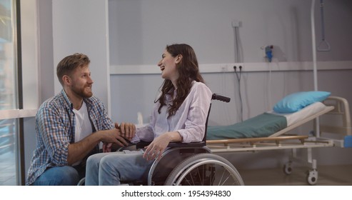 Young Man Bending Near Wife Sitting In Wheelchair In Hospital Ward. Sick Female Patient In Wheelchair With Husband Supporting Her At Rehabilitation Center Room