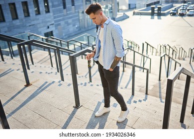 A young man with a black backpack standing on a city street holding sunglasses in his hands.