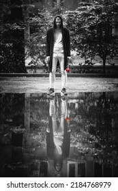 Young Man In The Black Cardigan With The Red Rose In The Hand Stands In Front Of The Puddle With The Reflection Of Himself. The Behind The Mirror Man. Sad Romance Concept.