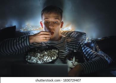 Young Man With Black Cat Watching A Movie Eating Popcorn On TV At Home. Movie Night. Relax,rest Watching A Horror Film Or Video On Screen. Background Lighting. Fun Scared Excited People On The Couch.