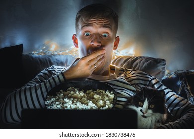 Young Man With Black Cat Watching A Movie Eating Popcorn On TV At Home. Movie Night. Relax,rest Watching A Horror Film Or Video On Screen. Background Lighting. Fun Scared Excited People On The Couch.