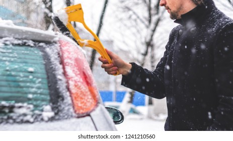 Young Man In Black Coat Cleans His Car With Yellow Brush During Snowfall. Winter Inclement Weather.