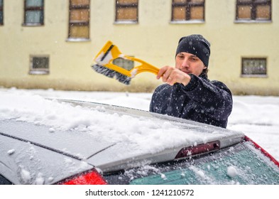 Young Man In Black Coat Cleans His Car With Yellow Brush During Snowfall. Winter Inclement Weather.