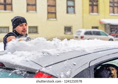 Young Man In Black Coat Cleans His Car With Yellow Brush During Snowfall. Winter Inclement Weather.