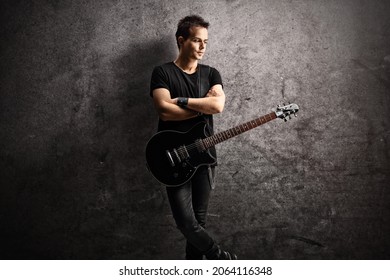 Young Man With A Black Electric Guitar On A Strap Leaning On A Gray Rugged Wall 