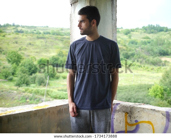 Young man with black hair and a beard in a gray\
T-shirt and gray Bermuda shorts stands on the terrace of an\
abandoned building, a serious expression on his face, left profile,\
looking to the right