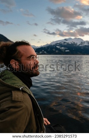 A young man in a black leather skirt and a military green jacket sitting on a pier next to a lake and looking with winter mountains background Stock photo © 