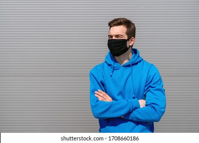 Young Man In A Black Mask With An Earring, Dressed In A Blue Hoodie, Black Jeans And Black Fitness Tracker, With A Serious Face And Cross Hands Look Into The Distance, Standing Against A Gray Textured