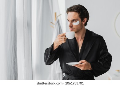 Young Man In Black Robe And Eye Patches Holding Coffee Cup In Bathroom