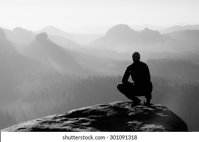 Young man in black sportswear is sitting on cliff's edge and looking to misty valley bellow