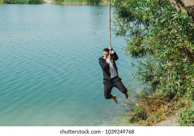 Young Man In Black Suit Is Jumping Into The Water. Wedding Photo For Groom Using The Bungee Over The Azure Blue Lake. Summer Fun Guy Photoshoot. Beautiful Nature And Trees Around.