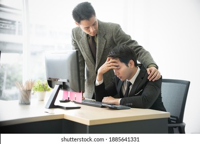 Young Man In Black Suit Sits At Work Desk In Front Of Computer Feel Sad, Headache, Depressed, Stressed While Standing Lawyer Lay Hand On His Shoulder To Support Him. Two Males Asian Homosexual Couple 