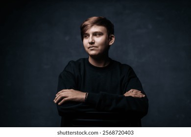 A young man in a black sweater sits with arms crossed, gazing thoughtfully into the distance. The dark background and soft lighting create a moody, introspective atmosphere. - Powered by Shutterstock