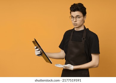 Young Man In A Black T-shirt And Apron In Work Gloves With A Tablet For Papers In His Hands Is Perplexed