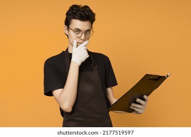 Young Man In A Black T-shirt And Apron In Work Gloves Thinks With A Tablet For Papers In His Hands