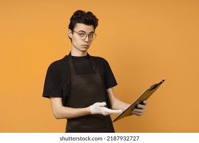 Young Man In A Black T-shirt And Apron In Work Gloves With A Tablet For Papers In His Hands Is Perplexed