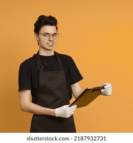 Young Man In A Black T-shirt And Apron In Work Gloves Holds A Tablet For Papers In His Hands