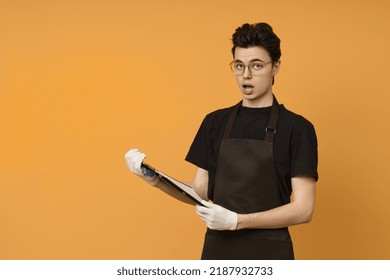 A Young Man In A Black T-shirt And Apron In Work Gloves Looks At The Camera In Surprise And Holds A Tablet For Papers In His Hands