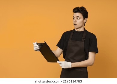 A Young Man In A Black T-shirt And Apron In Work Gloves Holds A Tablet For Papers In His Hands And Looks In Surprise At The Copy Space